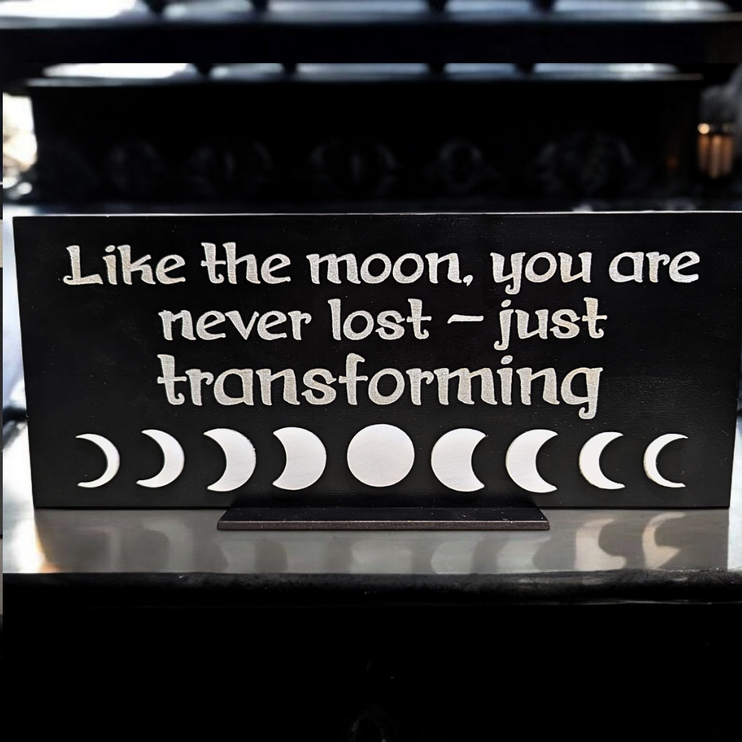 A black sign with a silver text that reads: 'Like the moon, you are never lost - just transforming.' Accompanied by a series of white acrylic moon phases below the text.