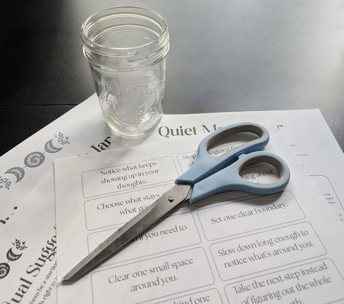 A clear glass mason jar sits on top of printed worksheets, with a pair of blue-handled scissors laid across them on a dark tabletop.