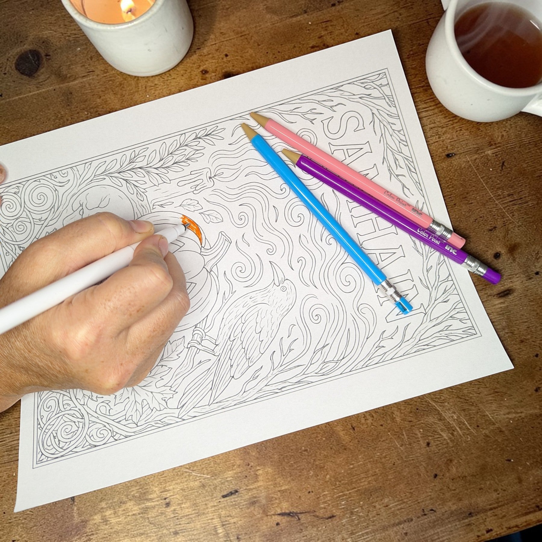 Person coloring a detailed design on paper with colored pencils, surrounded by a candle and cup of tea on a wooden table.